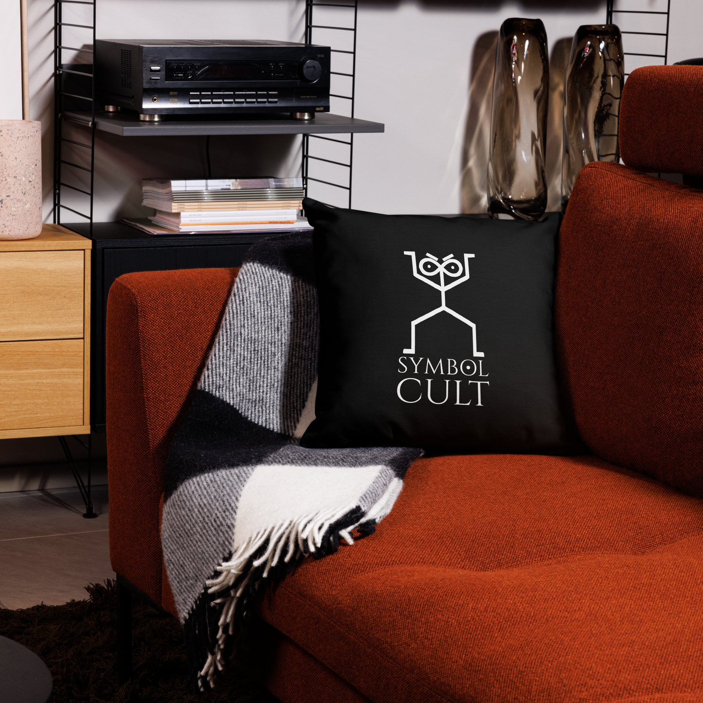 Symbol Cult Logo on a pillow the logo looks like an archic cave drawing of a humanoid figure with an infinity symbol for its head and dots in the symbol for pupils. it also has furrowed eyebrows that look to judge. below are the words Symbol Cult and the O in symbol is the alchemical symbol for the sun. all in white on black