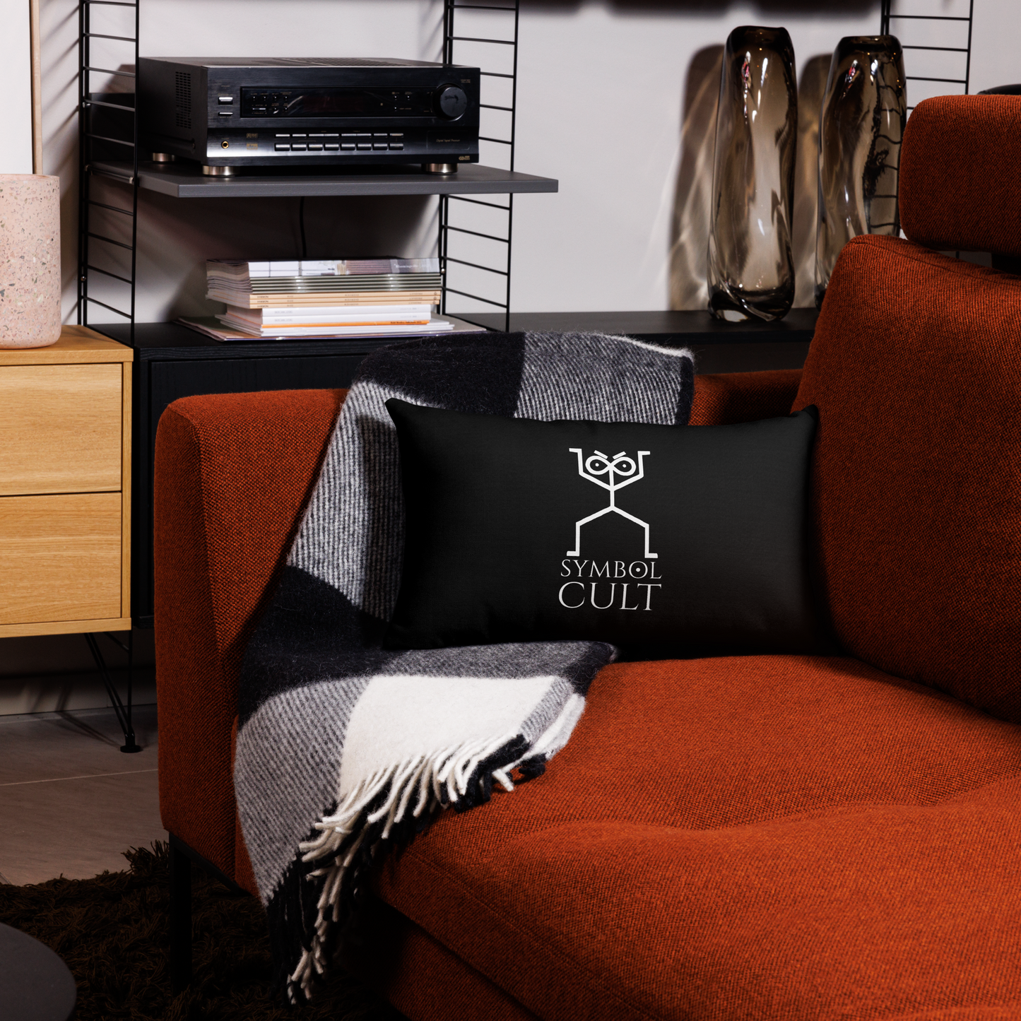 Symbol Cult Logo on a pillow the logo looks like an archic cave drawing of a humanoid figure with an infinity symbol for its head and dots in the symbol for pupils. it also has furrowed eyebrows that look to judge. below are the words Symbol Cult and the O in symbol is the alchemical symbol for the sun. all in white on black